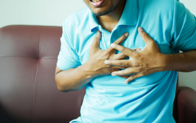 Person wearing a blue polo clutching their chest, seated on a couch, suggesting chest pain or distress in an indoor setting.