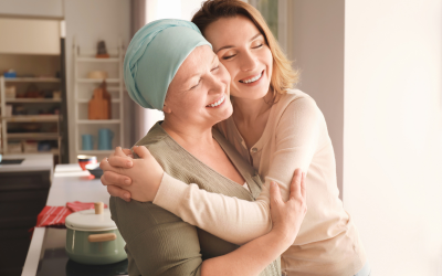 Caregiver hugs an elderly woman wearing a headscarf in a bright kitchen, both smiling warmly.