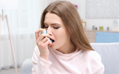 Woman seated indoors using a blue inhaler to relieve breathing difficulties in a bright living room.
